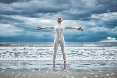 Happy hairless girl with alopecia in white futuristic suit stands with spread arms on beach bathed by ocean waves, performance of bald strong female artist symbolizing self acceptance clipart