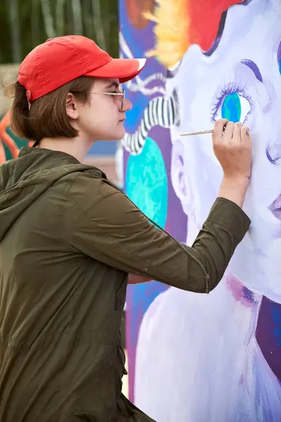 stock image Female painter in red cap draws surreal picture with paintbrush on canvas for outdoor street exhibition, close up side view of female artist apply brushstrokes to canvas, symphony of art creativity