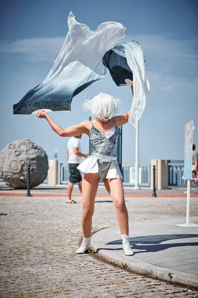 stock image Young sexy girl in space silver micro skirt dancing with performance fan waving gracefully, female outdoor dance on seaside promenade creating outdoor spectacle harmonizes with rhythm of sea waves