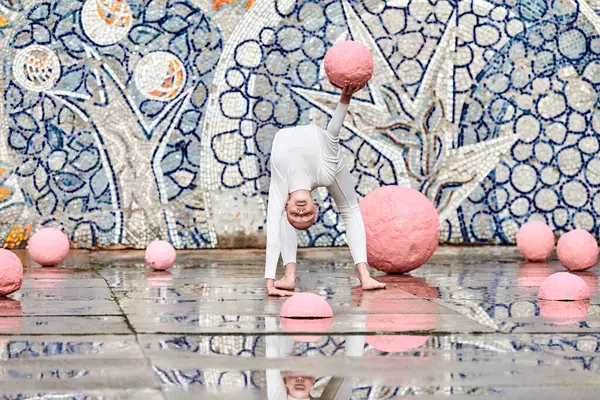 stock image Outdoor dance of young ballerina girl with alopecia in white futuristic suit with plastic and flexible movements among pink spheres on abstract mosaic Soviet background, symbolizes self expression