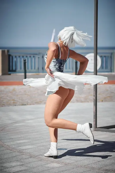 stock image Young sexy girl in space silver micro skirt dancing with white silk scarf waving gracefully, female outdoor dance performance on seaside promenade creating an arousing outdoor spectacle