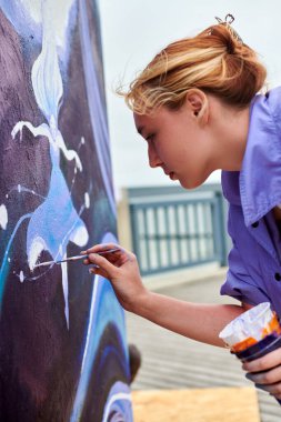 Kadın ressam, açık hava sokak sergisi için boya fırçasıyla resim çiziyor, kadın ressamın yan görüntüsünü kapatıyor fırça darbelerini tuvale uyguluyor, sanat yaratıcılığı senfonisi