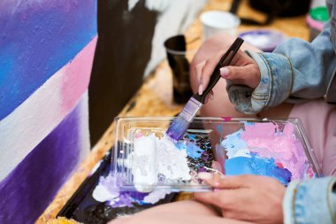 Kadın ressam el fırçasını açık hava sergisi için canlı resim paletine daldırıyor, elinde fırça tutan kadın ressamın görüntüsünü kapatıyor.