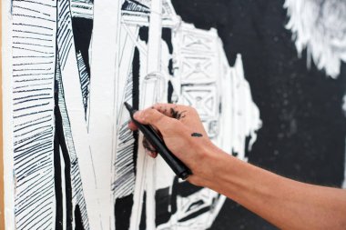 Genç erkek ressam, açık hava sokak sergisi için siyah keçeli kalemle resim çiziyor. Sadece siyah renk ve dışa dönük fırça darbeleriyle görsel bir manzara kullanıyor.