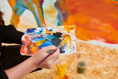 Kadın ressam el fırçasını açık hava sergisi için canlı resim paletine daldırıyor, elinde fırça tutan kadın ressamın görüntüsünü kapatıyor.