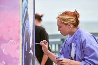Kadın ressam, açık hava sokak sergisi için boya fırçasıyla resim çiziyor, kadın ressamın yan görüntüsünü kapatıyor fırça darbelerini tuvale uyguluyor, sanat yaratıcılığı senfonisi