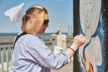 Güneş gözlüklü genç kadın ressam, açık hava sokak sergisi için boya fırçasıyla resim yapıyor. Parlak güneşli bir günde canlı sanat eserleri yaratmaya kendini kaptırmış kadın ressam.