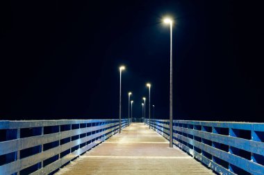 İskele boyunca gece karanlığına giden lambalar, denizin üzerindeki sakin, soğuk kış gecesini, sakin gece gökyüzünde minimalist rıhtımın geometrisini, insanlar olmadan açılacak olan limanı simgeler.
