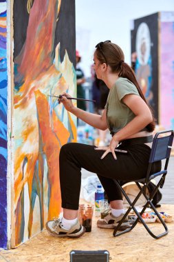 Çekici kadın ressam, açık hava sokak sergisi için sandalyede oturan boya fırçasıyla resim çiziyor, kadın ressamın dış mekâna hayat aşılamasının yan görüntüsü