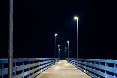 İskele boyunca gece karanlığına giden lambalar, denizin üzerindeki sakin, soğuk kış gecesini, sakin gece gökyüzünde minimalist rıhtımın geometrisini, insanlar olmadan açılacak olan limanı simgeler.