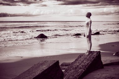 Kılsız, beyaz fütüristik takım elbiseli genç kızın tam boy portresi Rock Sea plajında duruyor, siyah beyaz portre, gerçek üstü sahne kel genç kız, koyu Magenta tonlu.