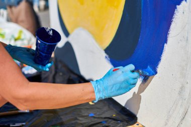 Kadın ressam eli, açık hava sokak sergisi için boya fırçasıyla canlı renkler kullanarak resim yapar, manzarayı kapatır