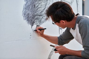Genç erkek ressam, açık hava sokak sergisi için siyah keçeli kalemle resim çiziyor. Sadece siyah renk ve dışa dönük fırça darbeleriyle görsel bir manzara kullanıyor.