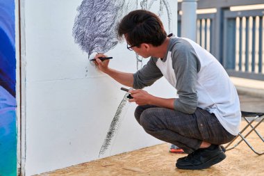 Genç erkek ressam, açık hava sokak sergisi için siyah keçeli kalemle resim çiziyor. Sadece siyah renk ve dışa dönük fırça darbeleriyle görsel bir manzara kullanıyor.