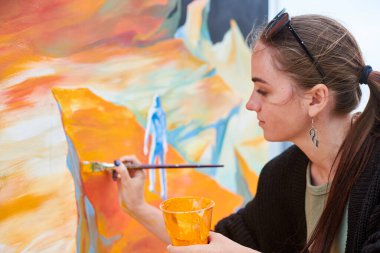 Çekici kadın ressam, açık hava sokak sergisi için boya fırçasıyla resim çiziyor, açık hava sanat alanına hayat veren kadın ressamın yan görüntüsünü kapatıyor.
