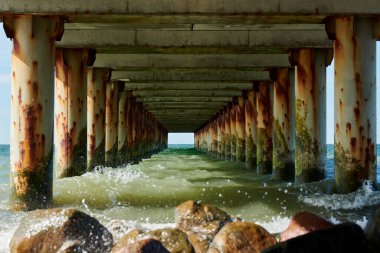 Eski deniz iskelesinin paslı sütunları iskelenin altından büyüleyici bir deniz manzarası yaratır. Huzursuz deniz dalgaları kayalara çarpar. Uyumlu ama güçlü kıyı senfonisini simgeler.