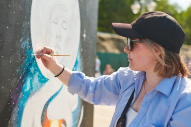 Siyah şapkalı ve güneş gözlüklü genç kadın ressam, açık hava sokak sergisi için boya fırçasıyla resim yapıyor. Parlak güneşli bir günde canlı sanat eserleri yaratmaya kendini kaptırmış kadın ressam.