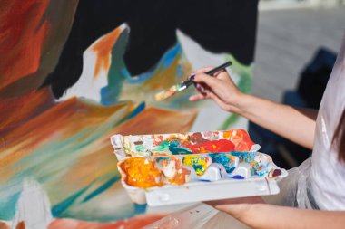 Kadın ressam eli, açık hava sokak sergisi için boya fırçasıyla canlı renkler kullanarak resim yapar, manzarayı kapatır