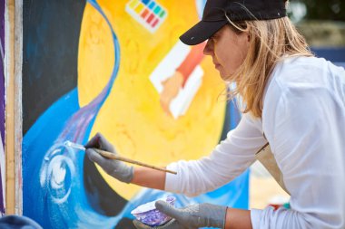 Siyah şapkalı kadın ressam, açık hava sergisi için tuval üzerine fırçayla resim çiziyor, kadın ressamın yan görüntüsünü kapatıyor fırça darbelerini tuvale uyguluyor, sanat yaratıcılığı senfonisi.