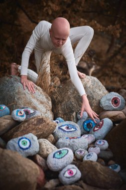 Young hairless girl with alopecia in white futuristic costume reaches hand for surreal landscape with lot of rocks with eyes, profound symbolism of embracing individuality in surreal world clipart