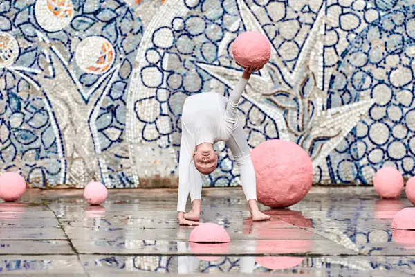 stock image Outdoor dance of young ballerina girl with alopecia in white futuristic suit with plastic and flexible movements among pink spheres on abstract mosaic Soviet background, symbolizes self expression