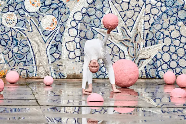 stock image Outdoor dance of young ballerina girl with alopecia in white futuristic suit with plastic and flexible movements among pink spheres on abstract mosaic Soviet background, symbolizes self expression