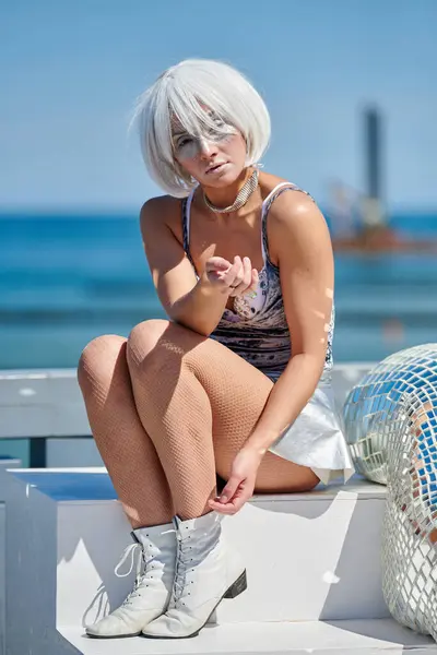 stock image Young sexy girl in space silver micro skirt and cosmic silver wig sits on white stairs next to an abstract space art object with lots of little mirrors, symbolizing tenderness and feminine sexuality