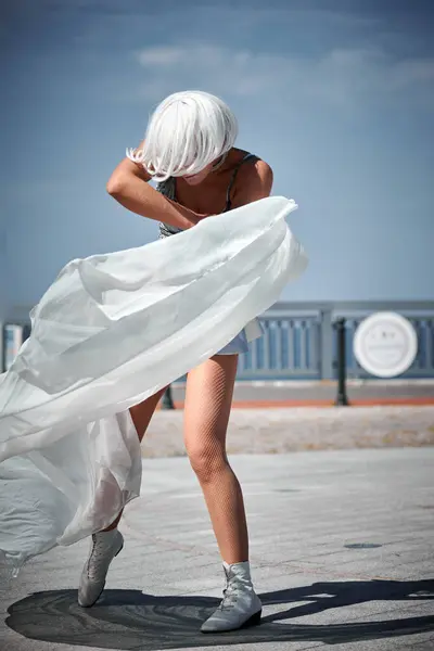 stock image Young sexy girl in space silver micro skirt dancing with white silk scarf waving gracefully, female outdoor dance performance on seaside promenade creating an arousing outdoor spectacle