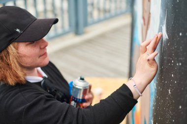 Female artist in black cap is painting picture with paint spray can spraying it onto canvas at outdoor street exhibition, side view of female art maker clipart