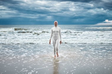 Beyaz fütüristik takım elbiseli, saçları dökülmemiş cesur bir kız kumlu sahilde soğuk denizden çıkar. Kel kadın sanatçının hayatın zorluklarını aşma ve kendini kabullenme metaforu.