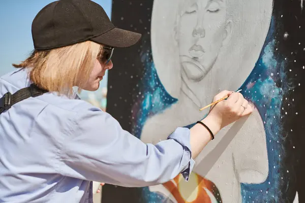 stock image Young female painter in black cap and sunglasses passionately paints picture with paintbrush for outdoor street exhibition, female artist engrossed in creating vibrant artwork at bright sunny day