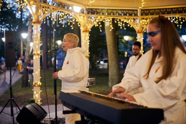Svetlogorsk, Russia - 13.08.2023 - Local pop rock music band performs in gazebo of city public park, side view of keyboardist to energetic outdoor performance in evening lights of park clipart