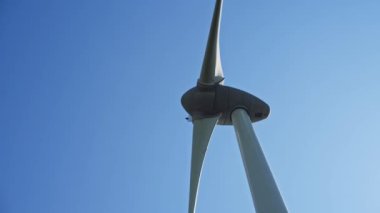 Kırmızı uçlu modern rüzgar türbini, canlı mavi gökyüzüne karşı dimdik duruyor. Dağılmış beyaz bulutlarla dolu, teknolojinin ve doğal ahengin bir karışımını yansıtıyor.