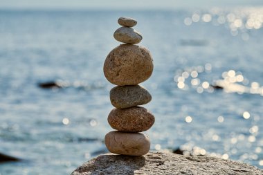 Sakin deniz kıyısında kayaların üzerinde dengelenmiş pürüzsüz taşlar yığını, denge ve sükuneti sembolize eder, barışçıl ve meditasyonel atmosferi çağrıştırır.