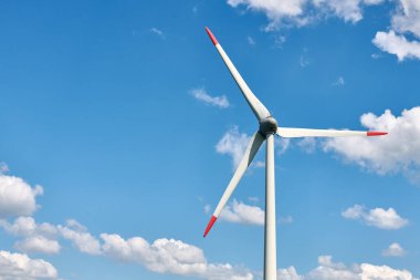 Kırmızı uçlu modern rüzgar türbini, canlı mavi gökyüzüne karşı dimdik duruyor. Dağılmış beyaz bulutlarla dolu, teknolojinin ve doğal ahengin bir karışımını yansıtıyor.
