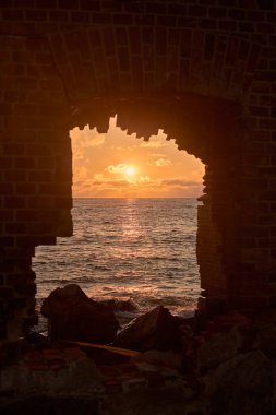 Yıpranmış tuğla pencere tarafından çerçevelenmiş büyüleyici sahil gün batımı, dinginlik ve nostalji uyandırıyor canlı renkler ve nazik dalgalar.