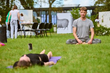 Yetişkin beyaz adam çimenler üzerinde derin derin meditasyon yapıyor, odaklanmış ve sakin. Kadın paspasın yanında yatıyor, rahat atmosferi ve hafif ışıklandırmasıyla arka planda park ediyor.