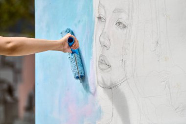 El, beyaz duvardaki genç kadın yüzünün çiziminin üzerine yumuşak mavi boya sürüyor. Doğal dış mekan, hafif ışıklandırma, sanatsal gelişim