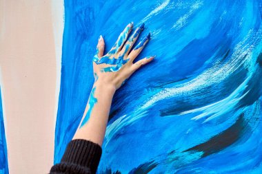 Yetişkin kadın eli, parlak mavi boyayla kaplı. Büyük tuvalde anlamlı darbeler yaratıyor. Kalın renkler ve dinamik hareket yaratıcılığı ve duyguyu iletir