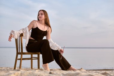 Genç bayan ressam kumlu sahilde tahta sandalyede oturuyor, elinde boya fırçası var. Huzurlu okyanus arkaplanı huzurlu bir ruh hali sağlar, sakin bir ilham uyandırır.