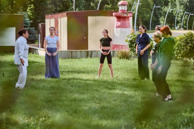 Çimenli bahçede qigong çalışan bir grup yetişkin. Güneşli gökyüzünün altında kadın öğretmen. Günlük kıyafetler, odaklanmış ifadeler, sakin enerji.