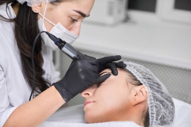 Yetenekli teknisyen, sakin salondaki genç bayana kalıcı kaş makyajı yapıyor. Siyah eldivenlerle, profesyonel aletler kullanılıyor, hassas ve rahatlatıcı ortamlarda bakım sağlanıyor.