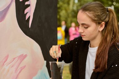 Açık havada resim yapan kahverengi saçlı genç bir kadın. Siyah kazak giyiyor, odaklanmış, canlı sanat yaratıyor. Arkaplanda ağaçlar ve bulanık şekiller yer alıyor