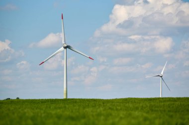 Rüzgar türbini, parlak mavi gökyüzünün altındaki yemyeşil buğday tarlasında kabarık beyaz bulutlarla dimdik ayakta duruyor.