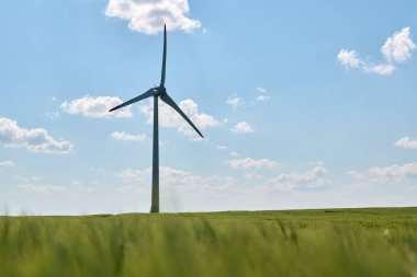 Rüzgar türbini, parlak mavi gökyüzünün altındaki yemyeşil buğday tarlasında kabarık beyaz bulutlarla dimdik ayakta duruyor.