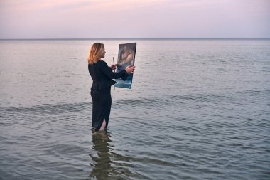 Dizlerine kadar okyanus suyuna batmış genç bir kadın, sakin deniz manzarasını yakalarken tuvaline konsantre oluyor. Sahne sükunet ve yaratıcılığı somutlaştırır, yumuşak akşam ışığı ile.