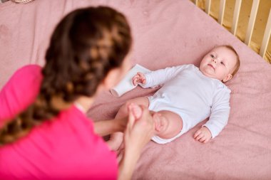 Kahverengi saçlı genç bayan yetişkin anne, küçük kızların ayağına krem sürüyor. Bebek, bebek odasında, yumuşak pembe battaniyenin üzerinde yatıyor. Bebek beyaz elbise giyiyor.