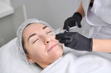 Kadın hasta klinik çevrede profesyonel olarak kozmetik enjeksiyondan geçiyor. Ayarlama sakin ve sterildir, estetik tedavilerde hassasiyet ve özen vurgulanır
