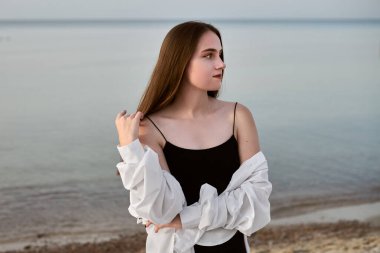 Siyah elbiseli ve beyaz gömlekli genç bir kadın sakin denizin kenarında derin derin duruyor. Rahat duruşu ve kapalı gözleri huzur ve huzur dolu sahili yansıtıyor.