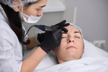 Güzellik uzmanı kuafördeki genç kadın kaşlarına kalıcı makyaj uygular. Odaklanma hassasiyet ve sanatsallık üzerinedir, sakin ortam deneyimi artırır.